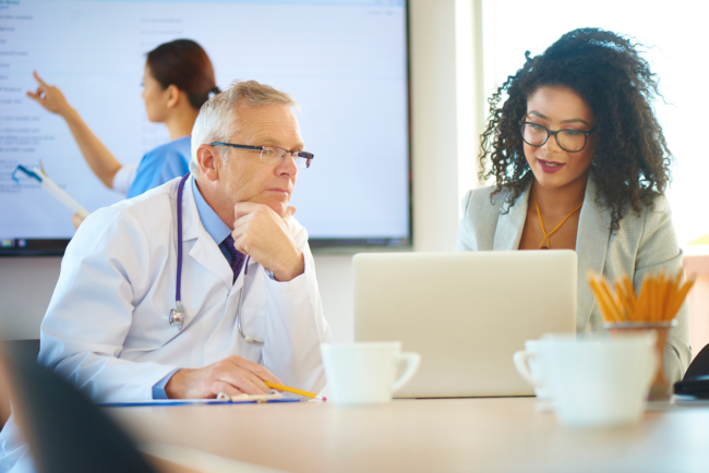 A doctor and an administrator review information on a laptop screen in a conference room.
